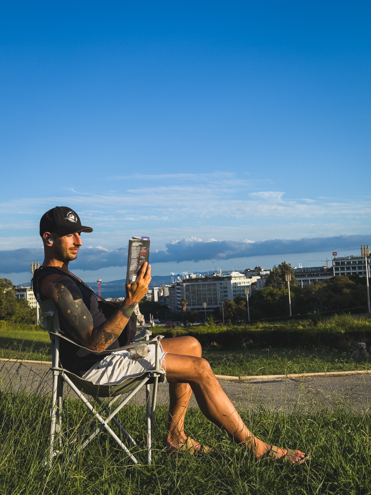 Simon Stawski reading in a portable chair on the grass at Parque Eduardo VII, with the Lisbon skyline and the Tagus behind him.
